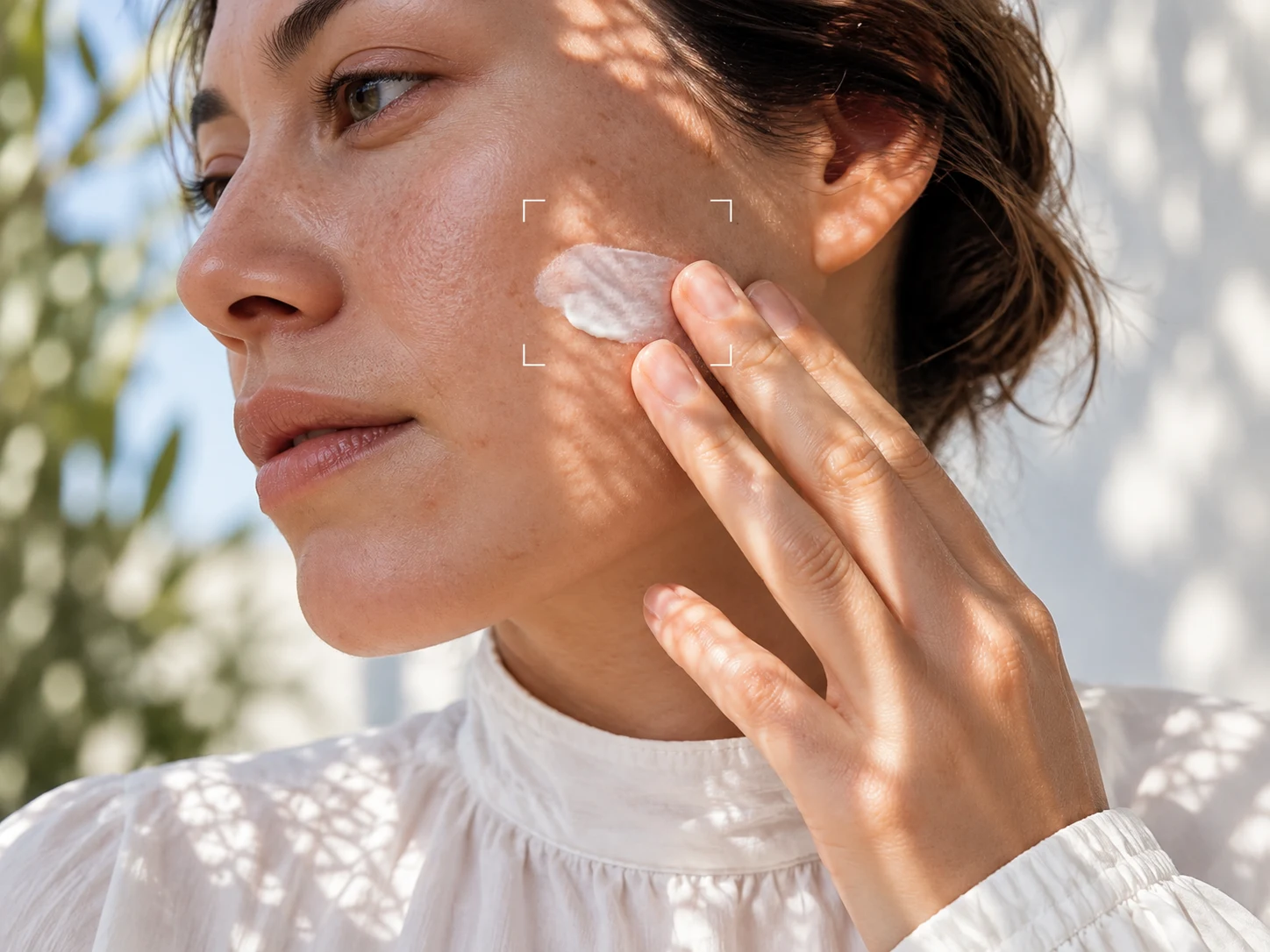 A woman in a white blouse applying sunscreen to her cheek in daylight.